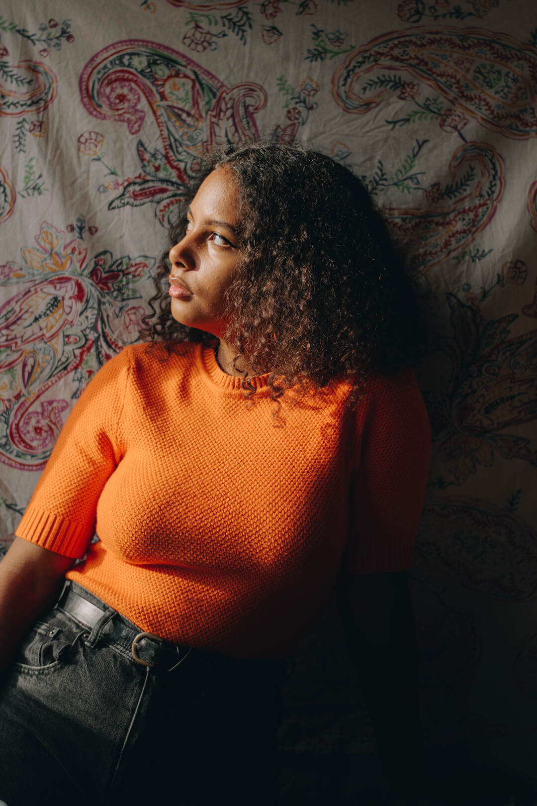 A young woman with curly hair wearing an orange sweater poses in a studio setting.