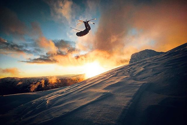 A skier performs a backflip against a sunset backdrop in a snowy mountain landscape.