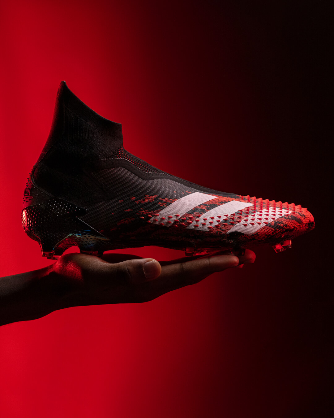 A hand holds an adidas Predator soccer shoe against a vibrant red backdrop in a studio setting.