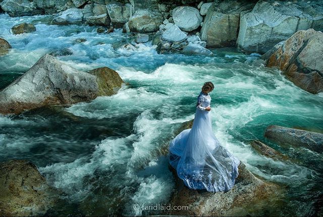 A bride in a white wedding dress stands on a rock in a rushing river.