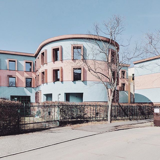 A modern pastel building with rounded architecture stands peacefully on a clear day, surrounded by greenery and a fence.