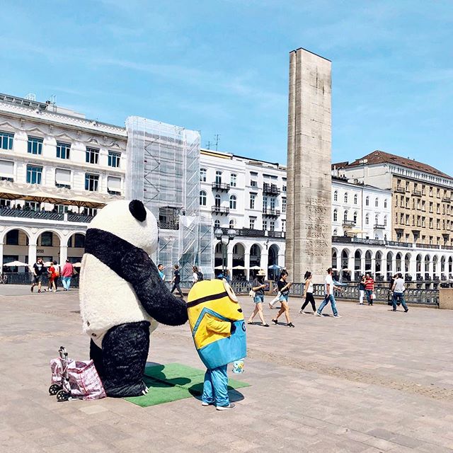 Two street performers dressed as a panda and a minion entertain passersby in a sunny city square.