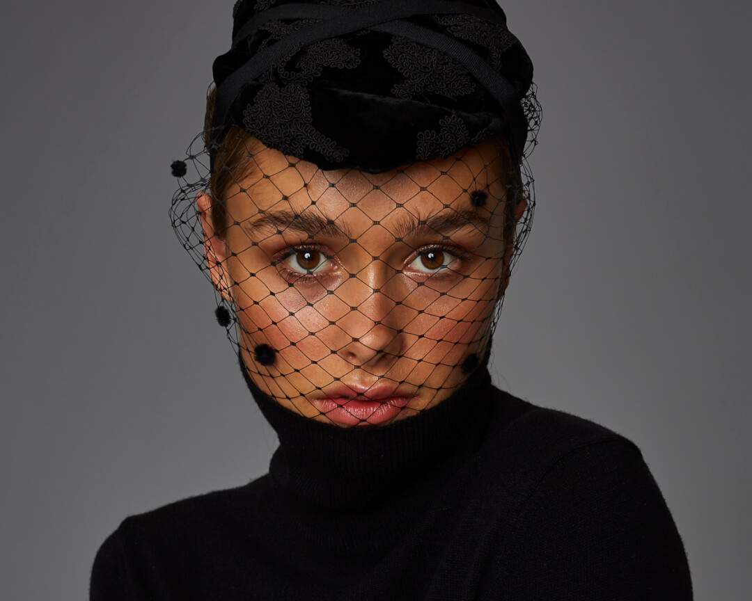 A woman with a black net veil and turtleneck poses for a fashion portrait.