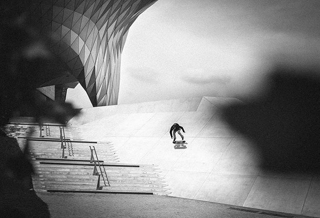 A skateboarder rides down a concrete incline next to modern architecture in a monochrome urban setting.