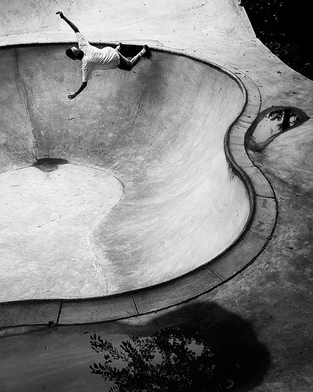 A skateboarder in a black and white photo is caught mid-air, riding the walls of a concrete skatepark bowl.