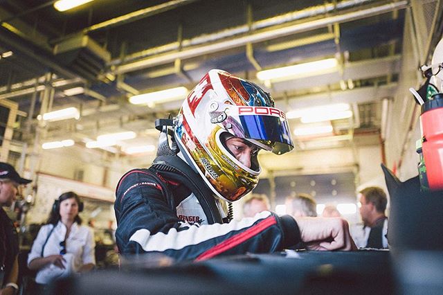 A racer wearing a helmet and racing suit is preparing for a race in the garage.