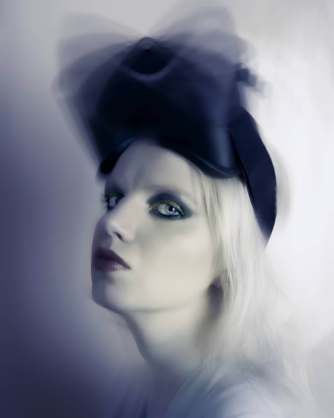 A woman with blonde hair and dramatic eye makeup poses with a unique hat in a fashion editorial style.