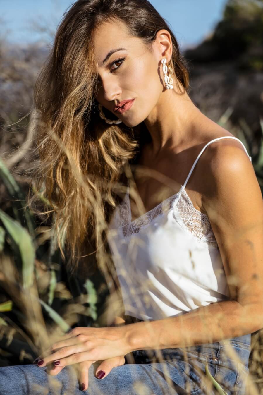 A beautiful brunette woman in a white top poses outdoors with soft, natural lighting for a serene editorial shot.