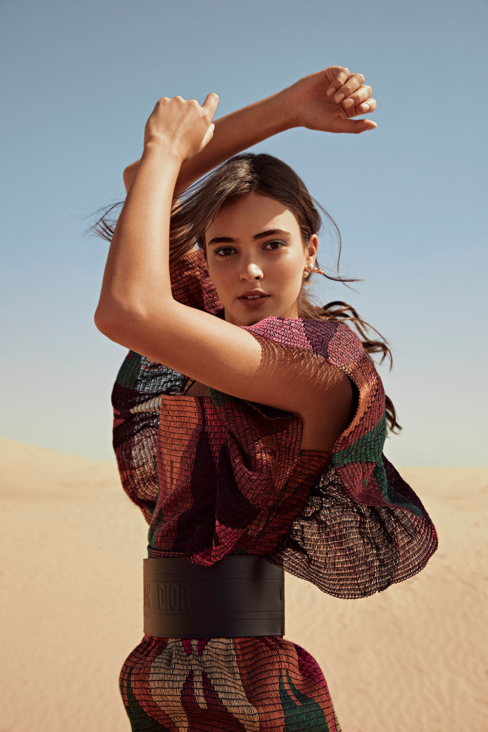 A confident woman poses in a colorful dress with a Dior belt against a desert backdrop for a fashion editorial.