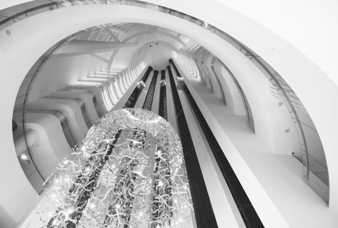 A monochrome low-angle shot of an atrium with a chandelier and elevator shafts conveys architectural sophistication.
