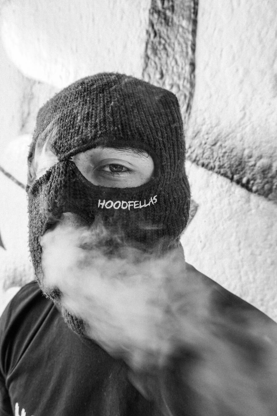 A person wearing a ski mask stands in front of a wall, smoke obscures the lower part of the face.