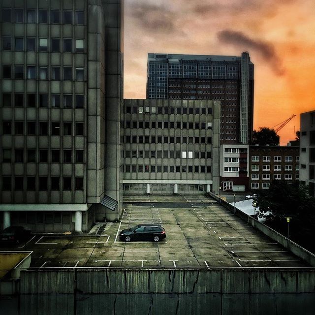 A car sits in a parking lot, framed by large buildings and a dramatic sunset sky.