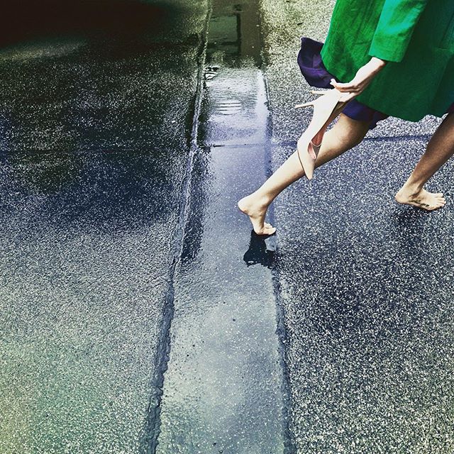 A barefoot woman in a green dress walks on a wet city street, holding her shoes.