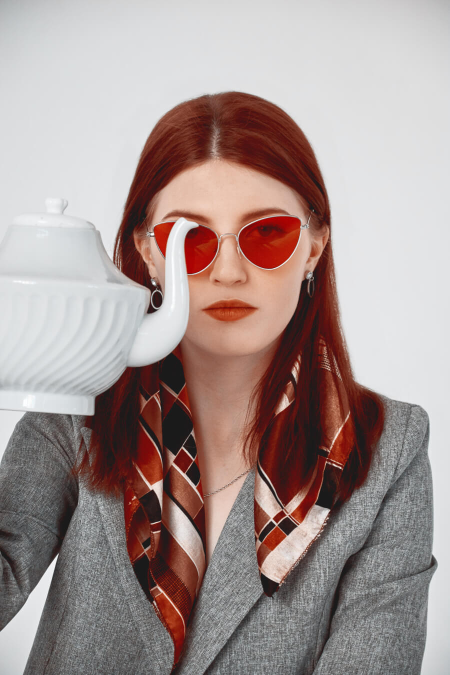 A stylish woman in red sunglasses and a blazer poses with a teapot for an editorial shot.