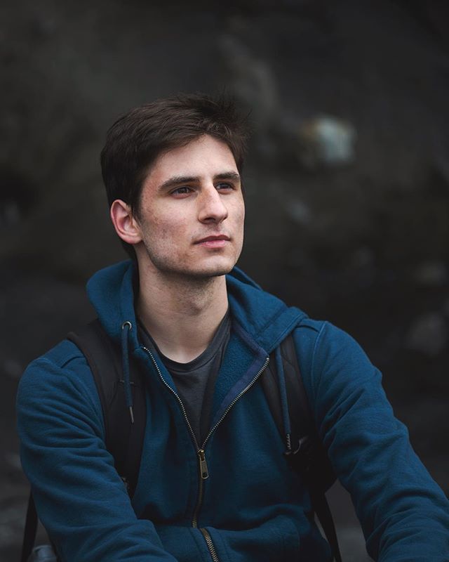 A man in a blue hoodie looks thoughtfully off-camera in a natural outdoor setting.