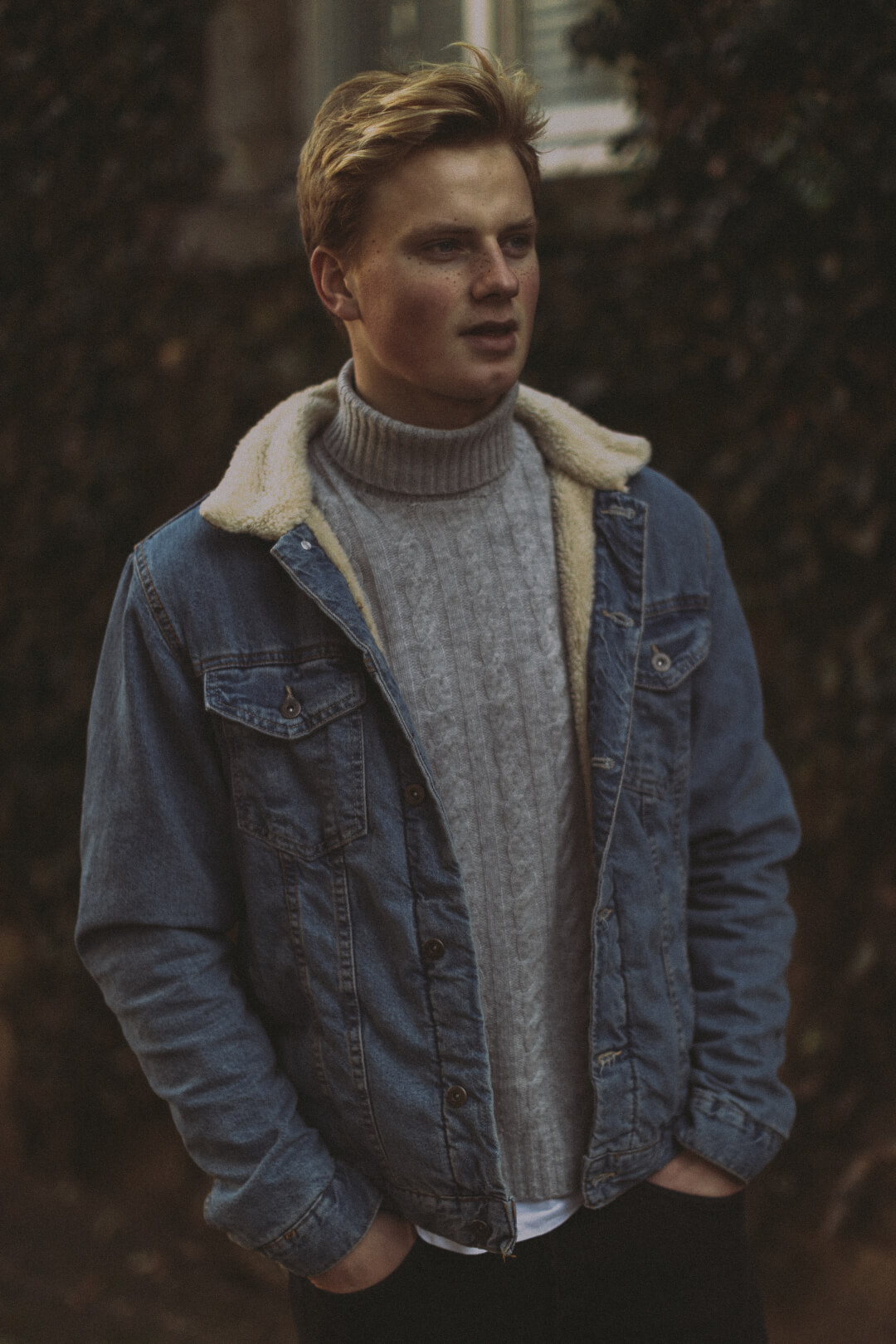 A blond man in a denim jacket and turtleneck sweater poses outdoors in a calm, casual setting.