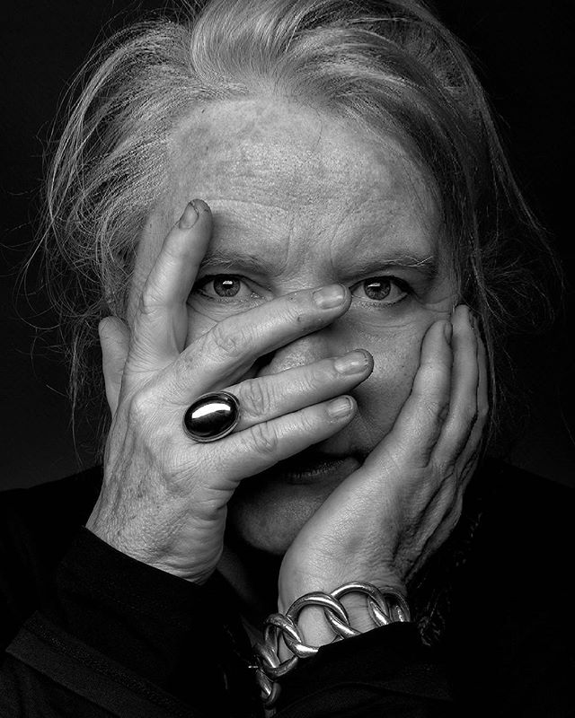 A monochrome portrait of a woman with silver hair, her face partially obscured by her hands and jewelry.