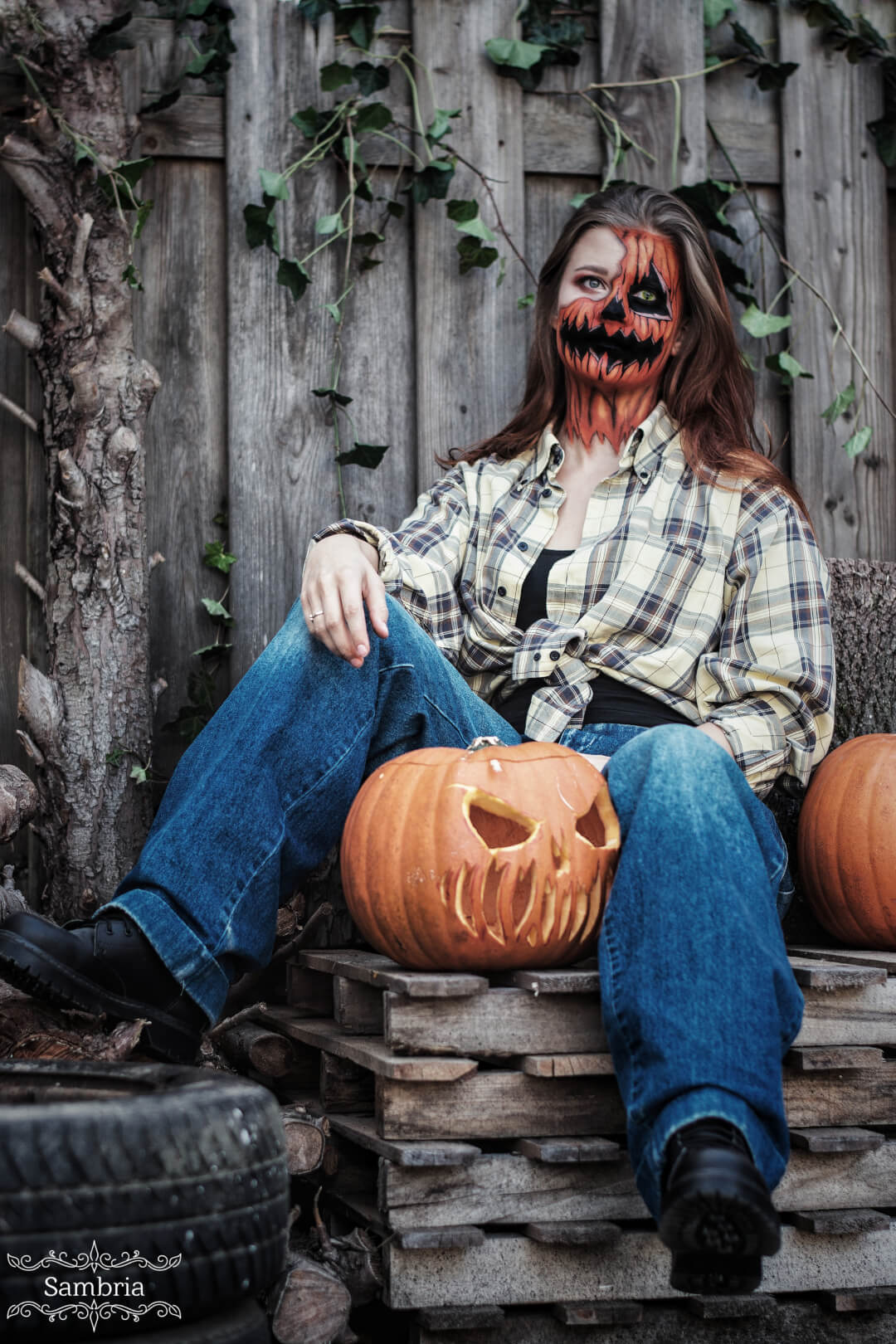 A woman with jack-o'-lantern makeup sits outdoors with a carved pumpkin for Halloween.