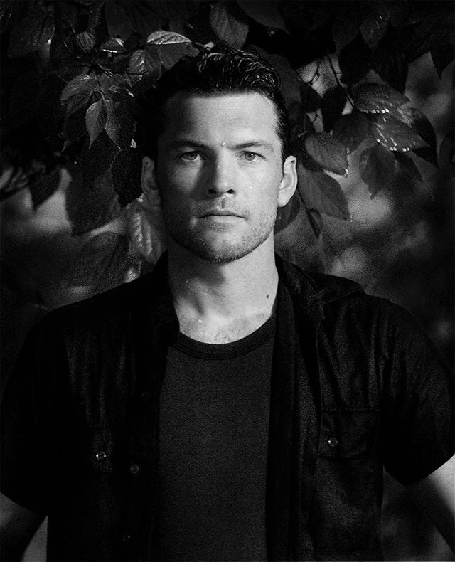 Black and white portrait of actor Sam Worthington with leaves in the background.