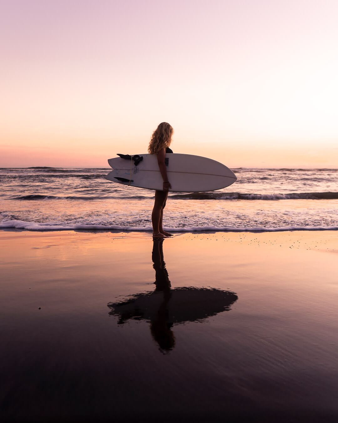 A woman with a surfboard stands on a beach at sunset, reflected in the wet sand.
