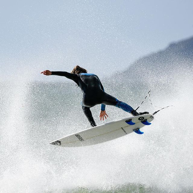 A surfer in a black wetsuit performs a trick mid-air on a white surfboard with blue fins over splashing ocean waves.
