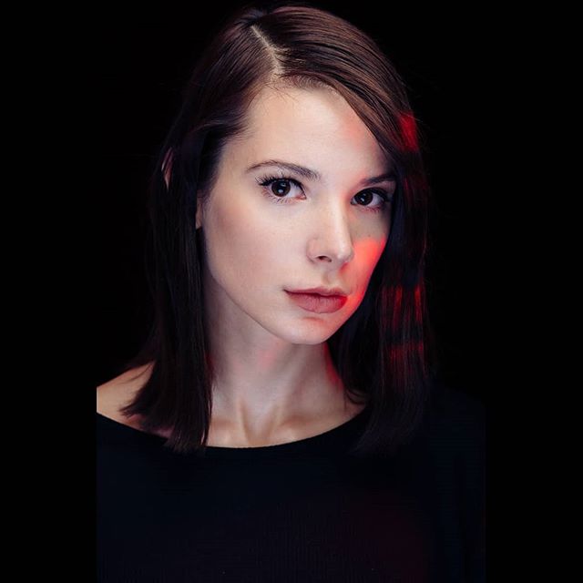 A brunette woman with fair skin poses in a studio with a dark background and red lighting.