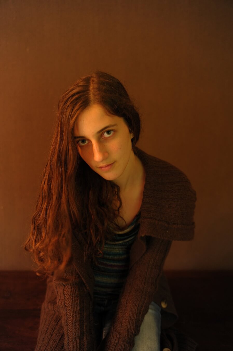 A young woman with long hair poses indoors with a thoughtful expression in soft, warm lighting.