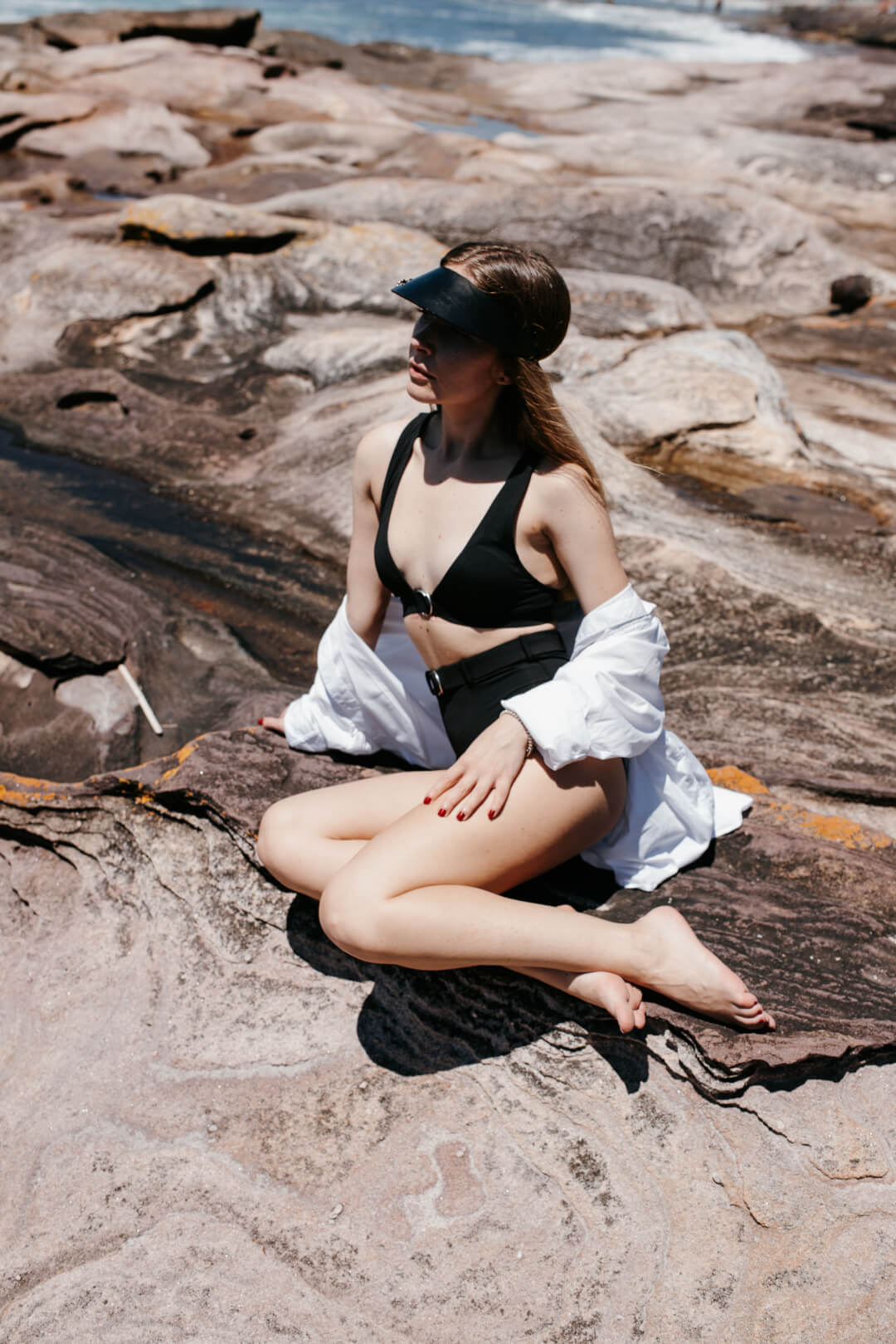 A woman in a black swimsuit and white shirt sits on rocks by the beach in the sunshine.