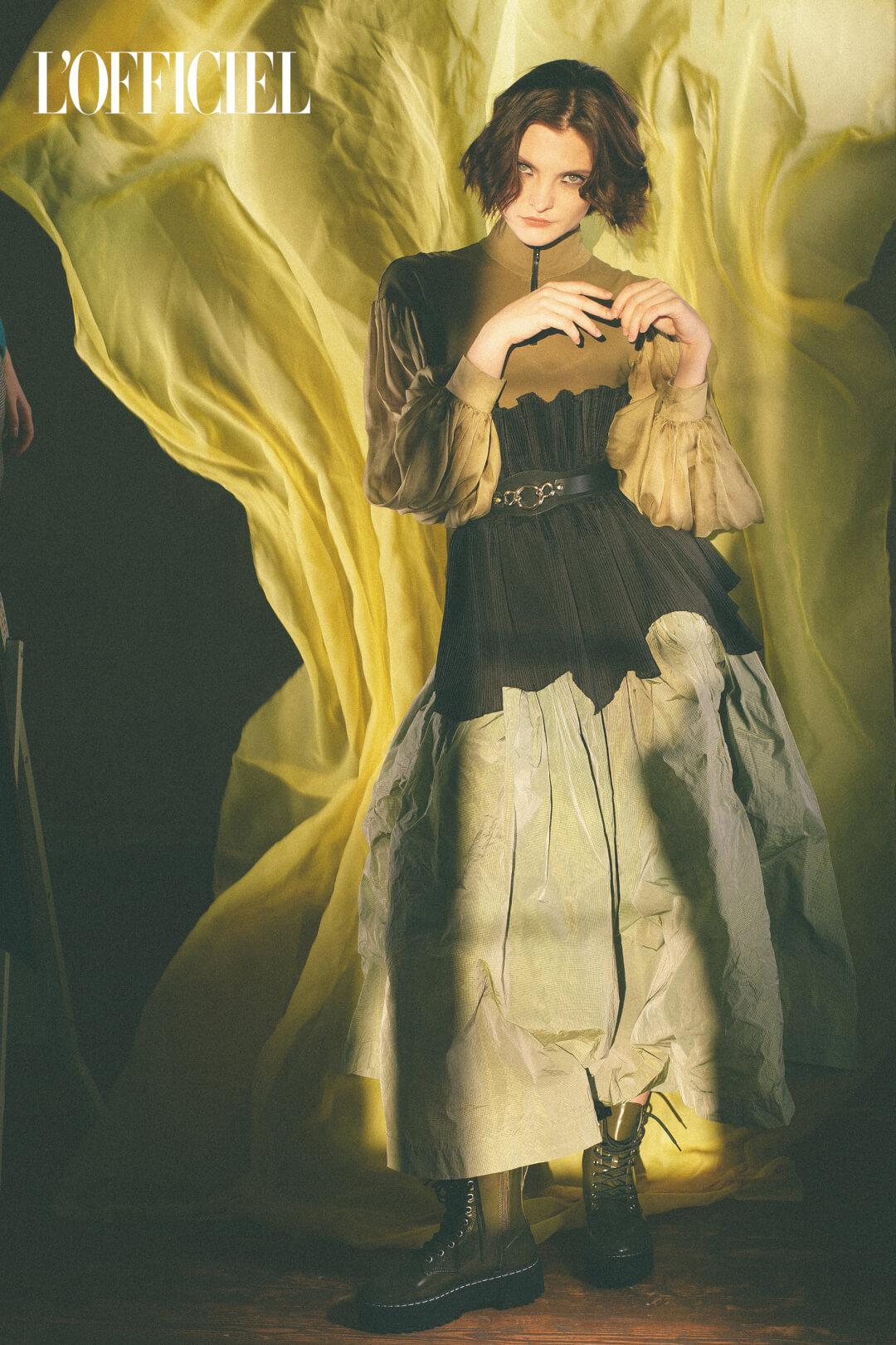 A fashion model poses in an olive green and black avant-garde dress with dramatic lighting for a high fashion editorial.