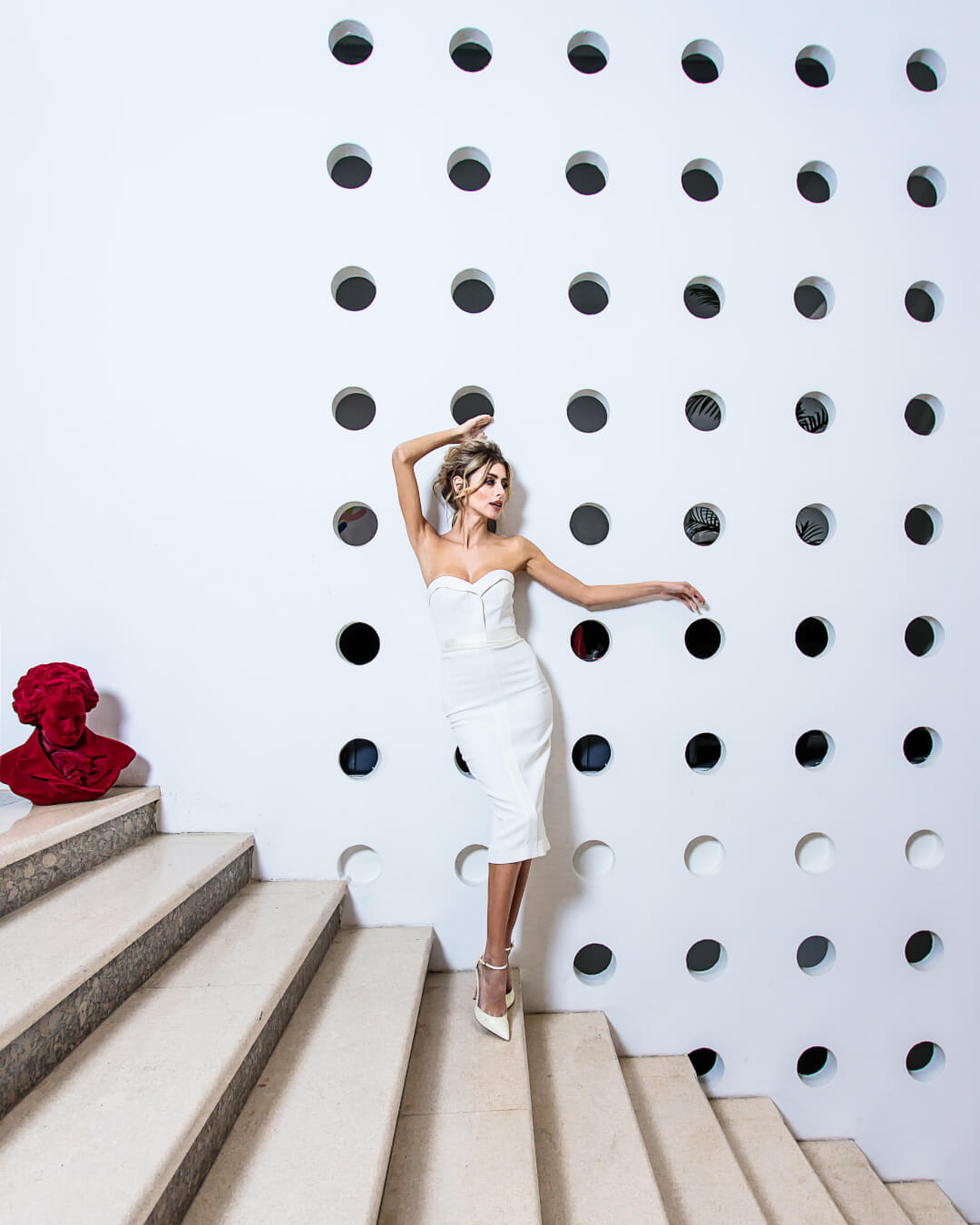A woman in a white dress poses on stairs next to a red bust, highlighting modern fashion against architectural backdrop.