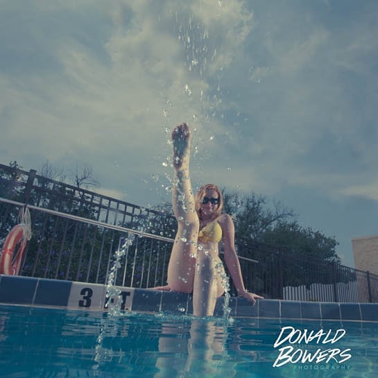 A woman in a swimsuit playfully kicks water in a swimming pool on a sunny day.
