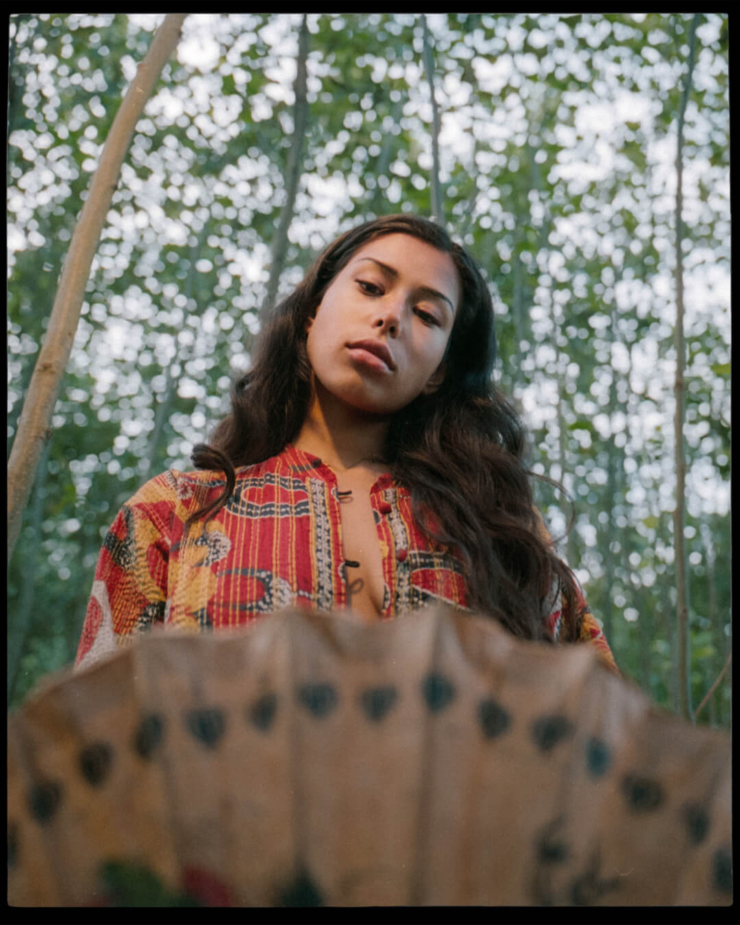 A young woman poses outdoors in a forest setting with dappled light and wearing a colorful patterned jacket.
