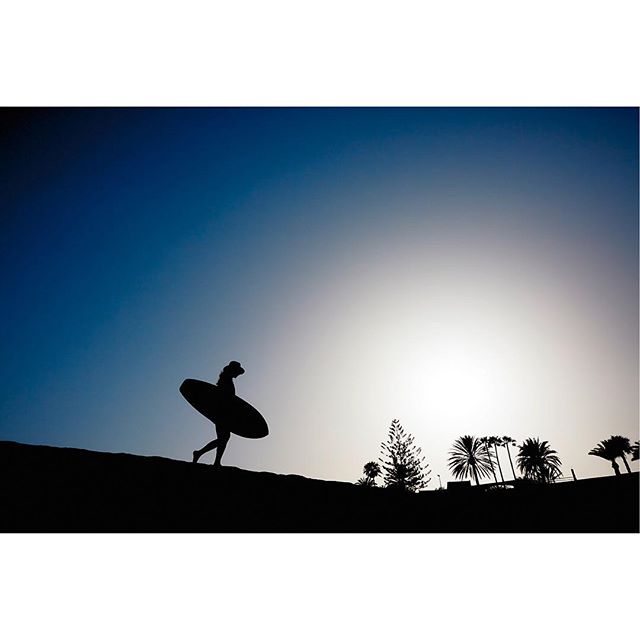 A silhouetted woman with a surfboard walks on a beach at sunset.