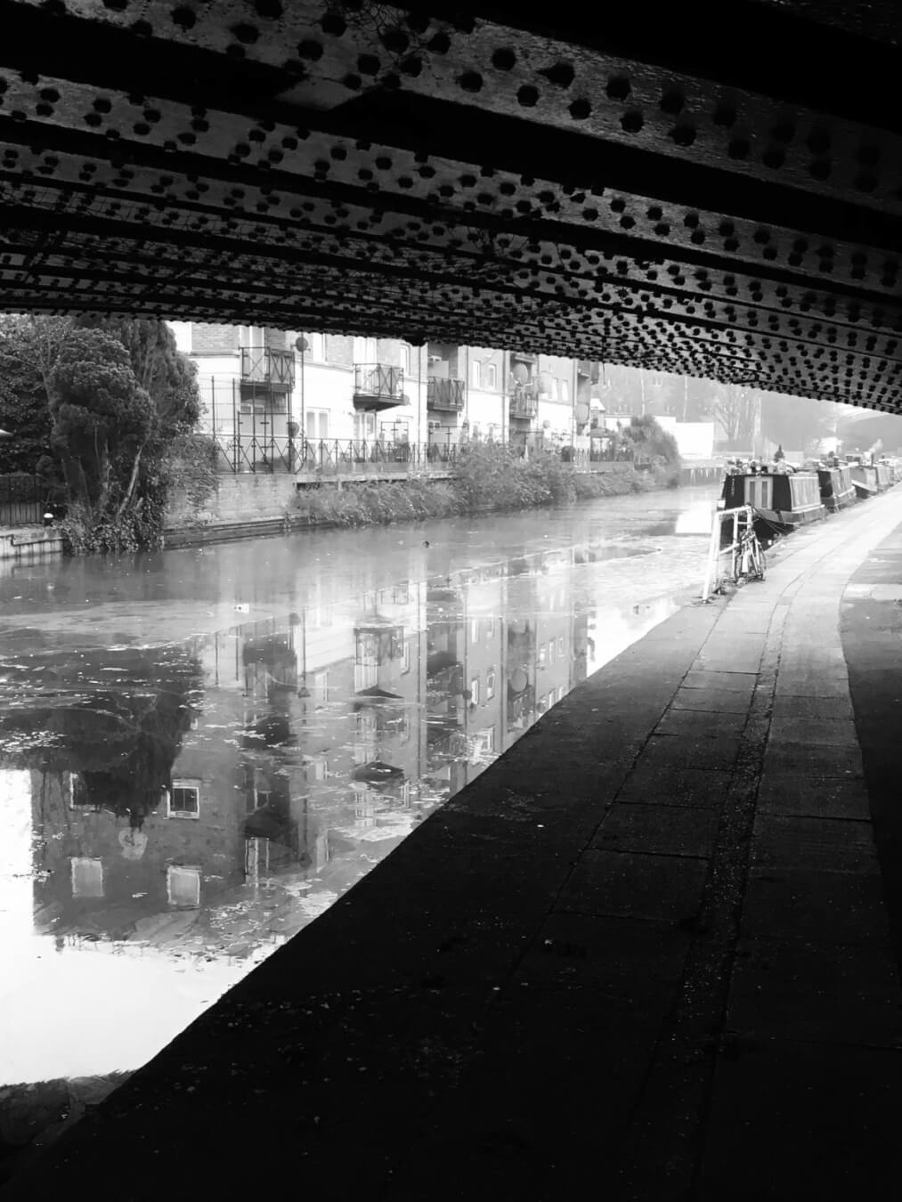 A serene black and white view under a bridge over a canal with buildings reflecting in the water.