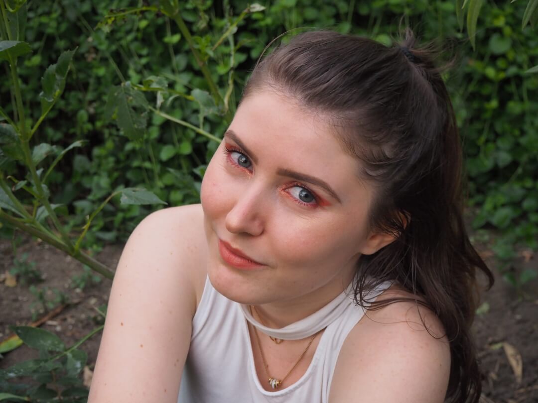 A young woman with red eyeshadow looks directly at the camera, enjoying a peaceful moment in a lush garden.