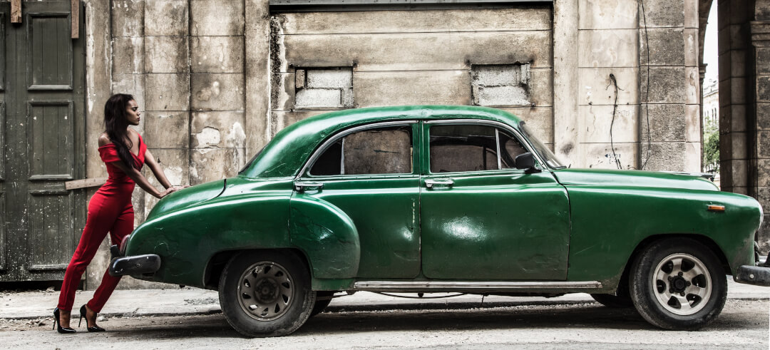 A woman in a red jumpsuit poses with a green vintage car against a weathered building, creating a fashion editorial scene.