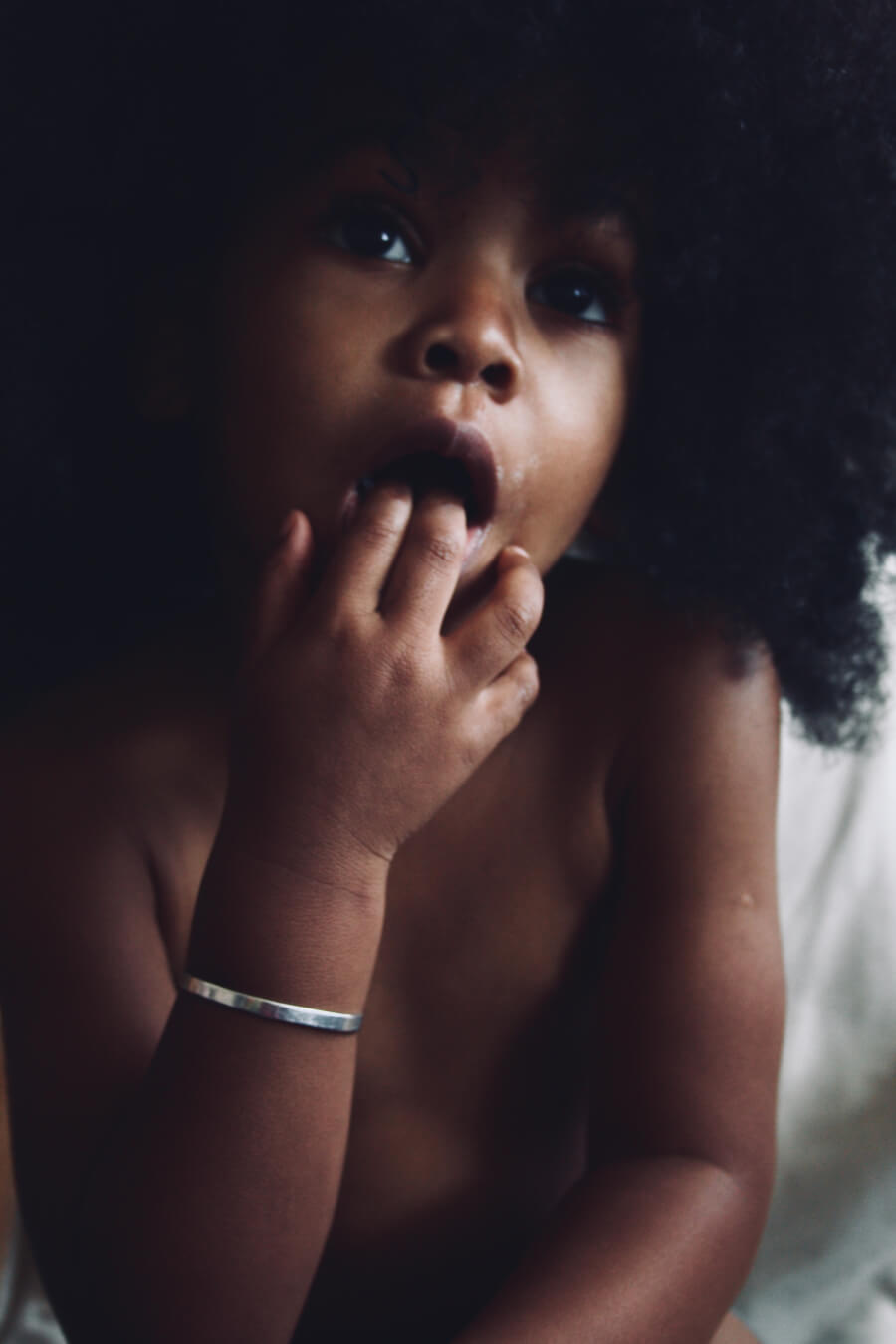 A cute child with an afro sits with fingers in their mouth, a soft, dreamy portrait.