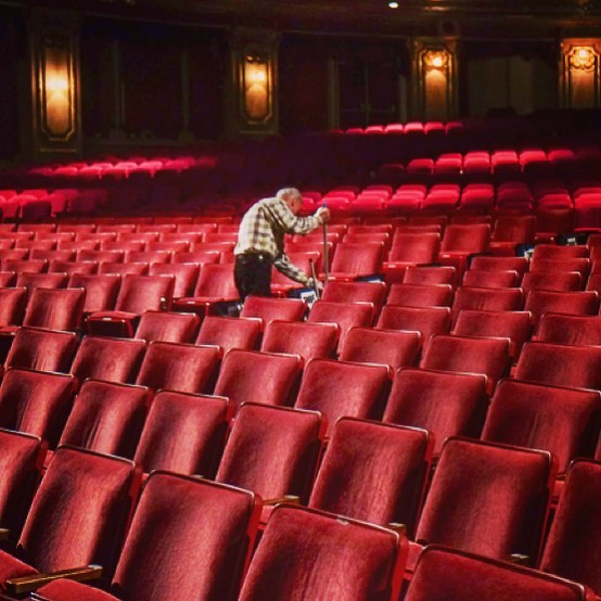 A man cleans between rows of red seats in a vintage theater auditorium.