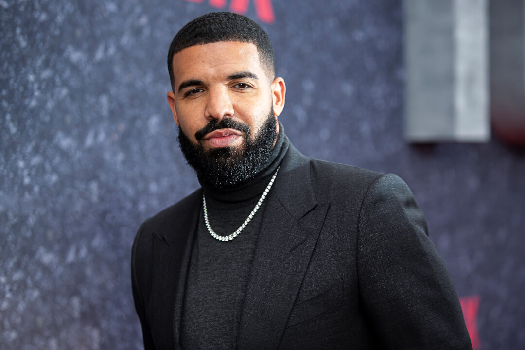 Drake poses on the red carpet wearing a black turtleneck and suit jacket with a diamond necklace.