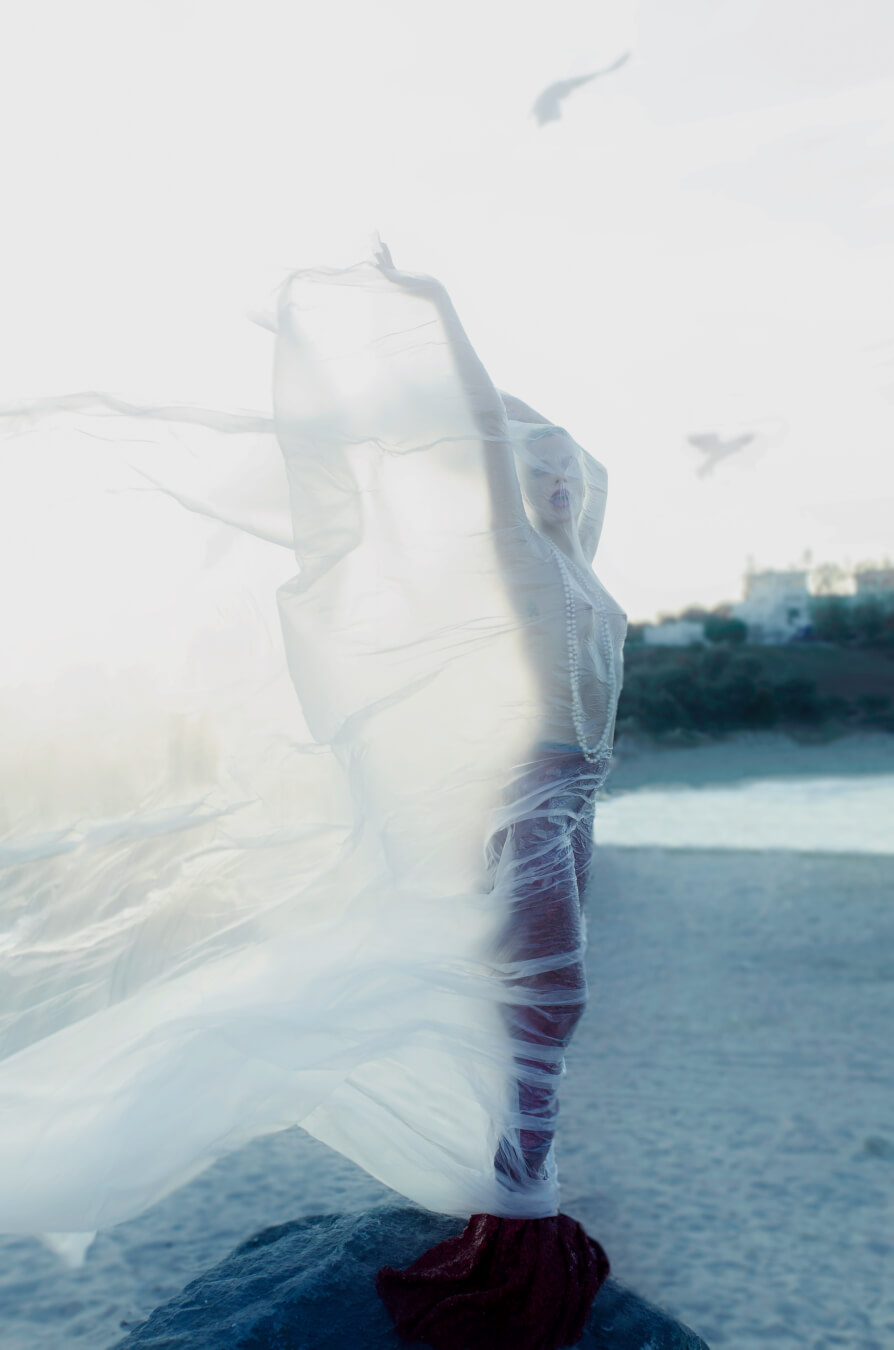 A woman draped in sheer fabric poses on a beach with a serene, dreamy quality, highlighting artistic nude fashion.