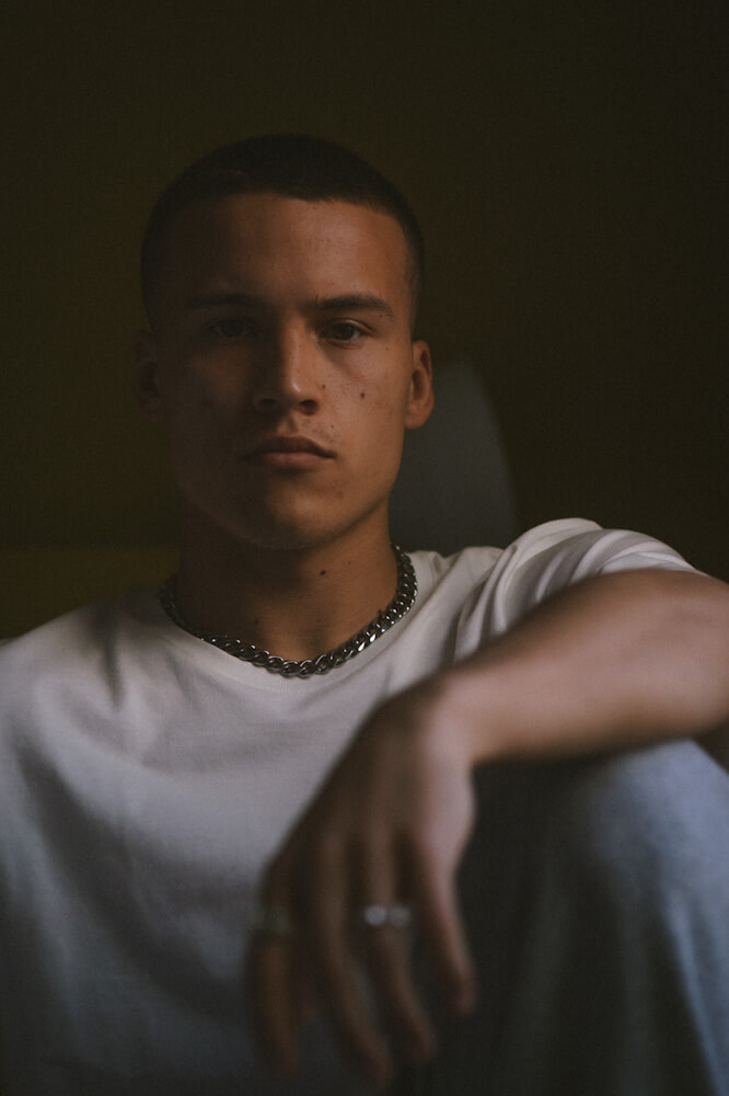 A young man poses indoors with a chain necklace, capturing an intimate portrait.