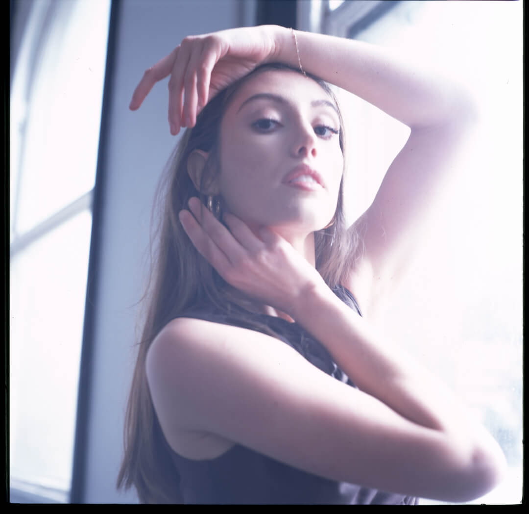 A young woman poses in soft window light, creating a sensual and dreamy portrait for editorial or advertising use.