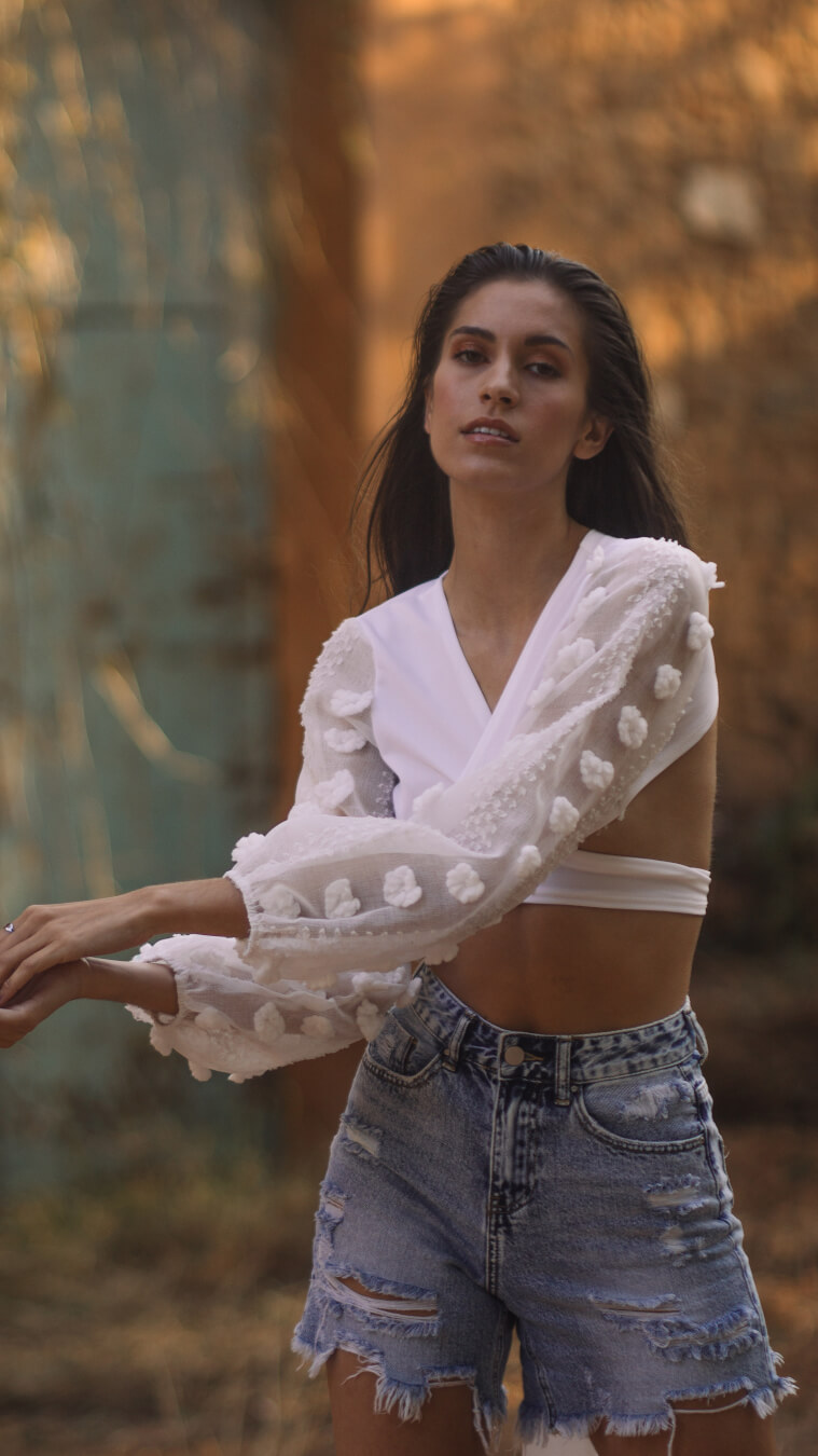 A brunette woman poses in a white crop top and distressed denim shorts, outdoors.