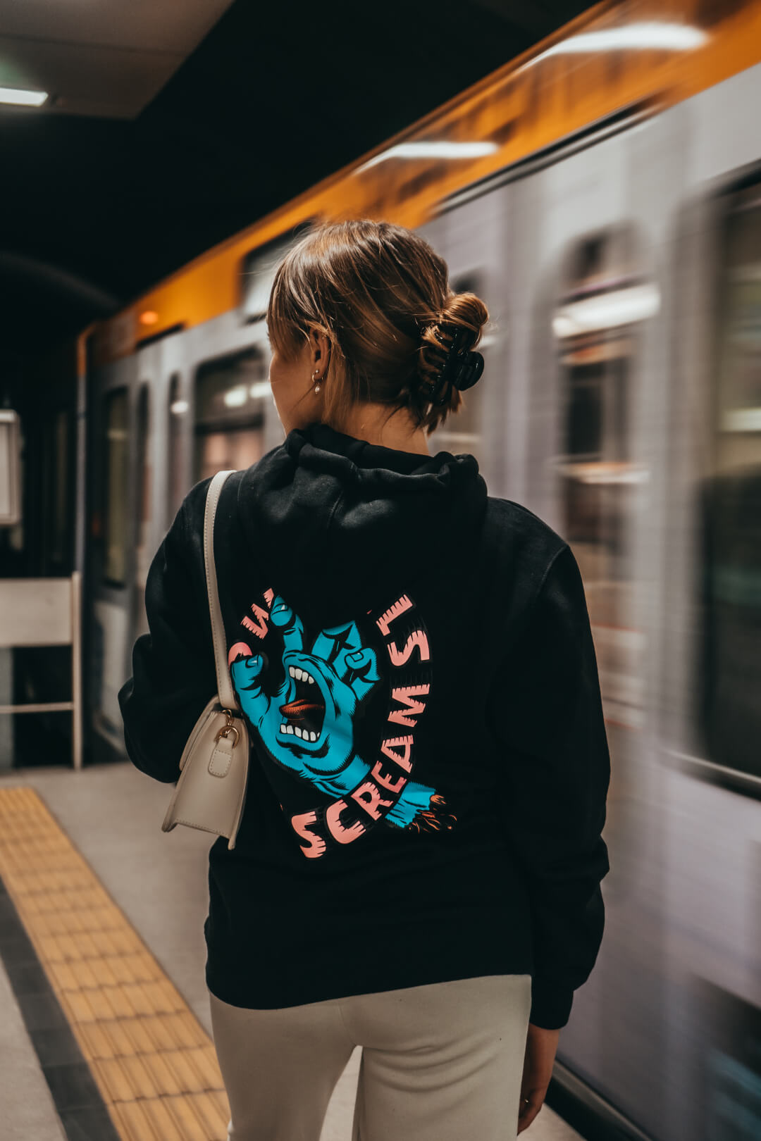 A woman waits at a train station with a train passing by, wearing a graphic hoodie.