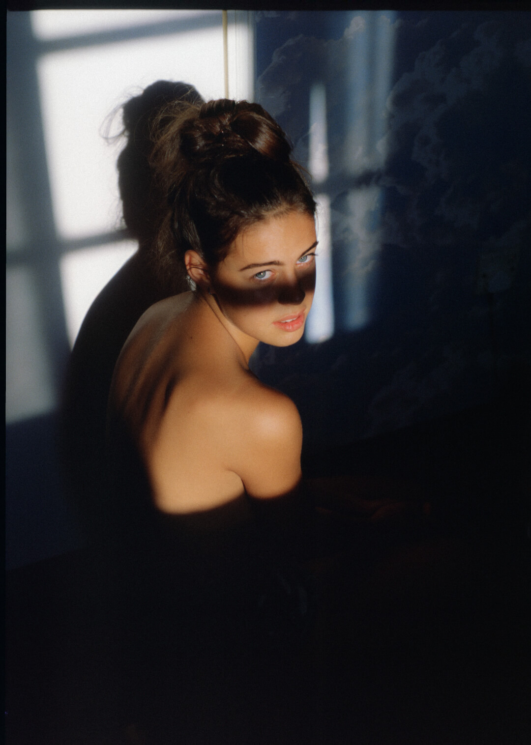 A young woman gazes over her shoulder in soft, dramatic window lighting.