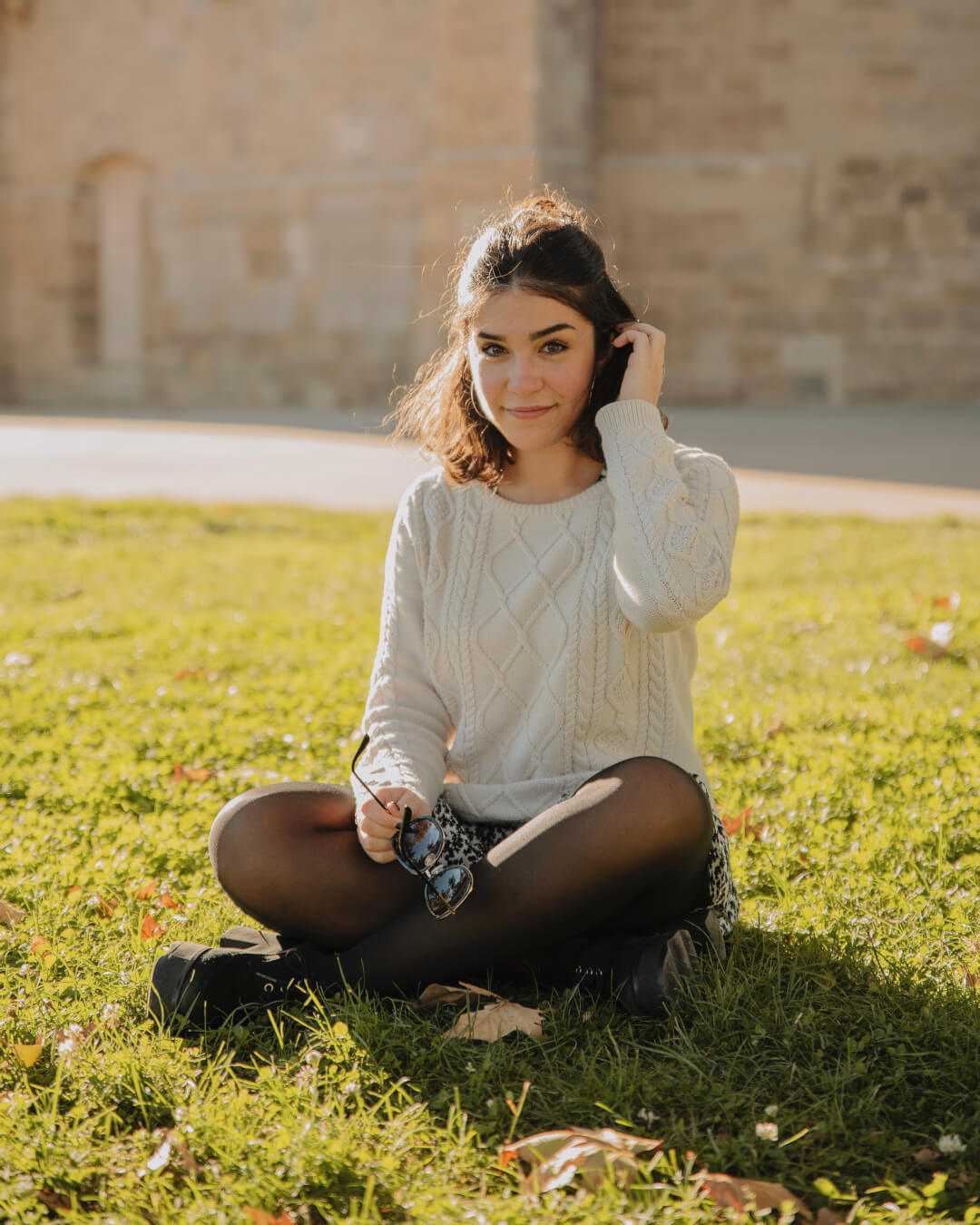A young woman sits cross-legged in the grass with sunglasses in her hand, enjoying a peaceful moment outdoors.