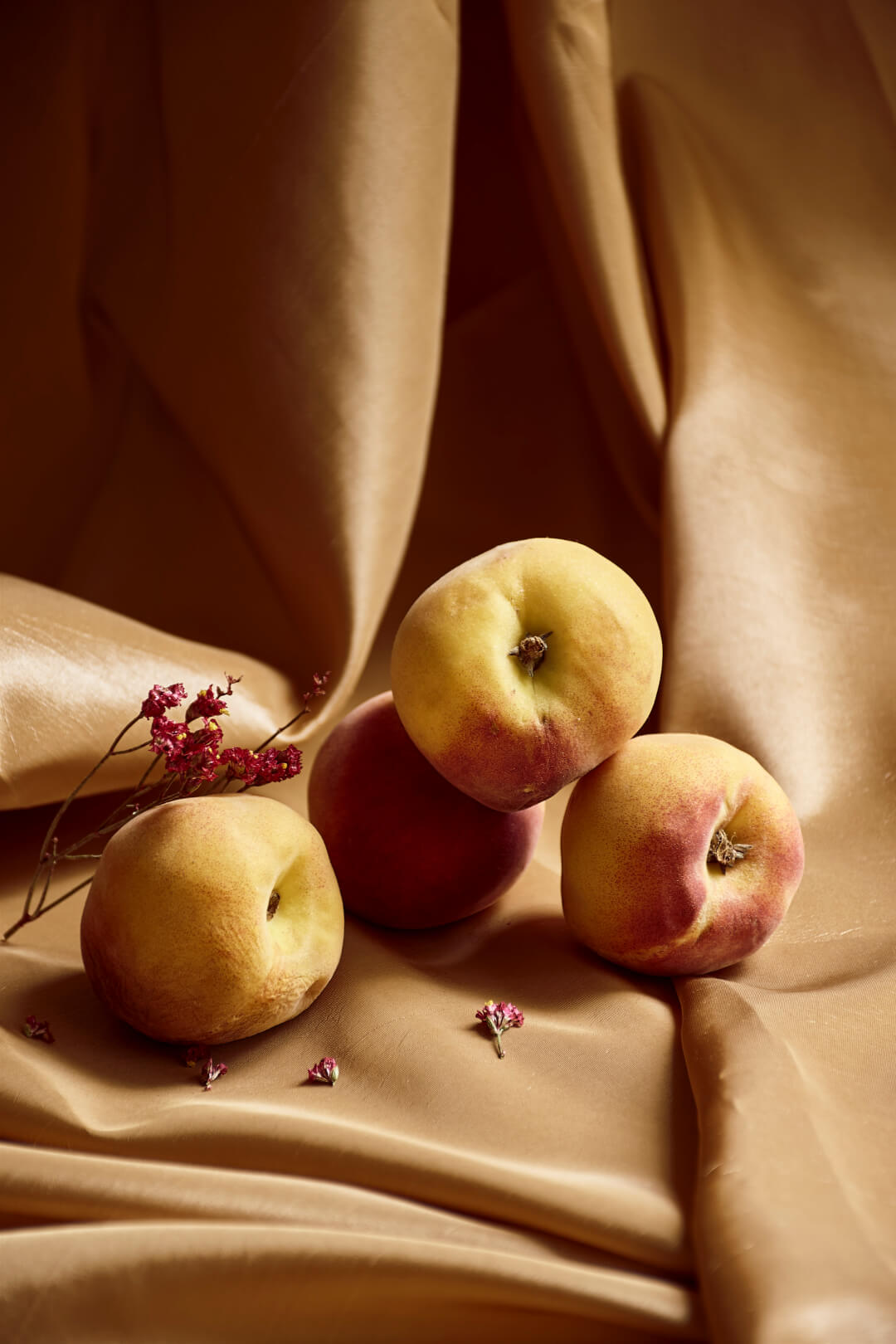 A still life featuring a collection of peaches with small red flowers against a draped golden fabric background.