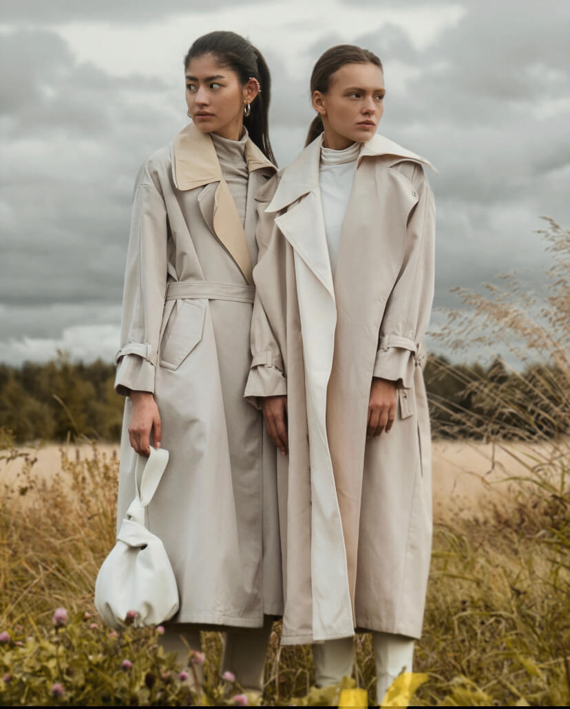 Two women model elegant beige trench coats against a cloudy sky in a field.