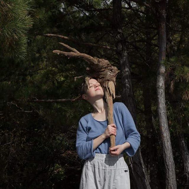 A woman embraces nature, holding driftwood in a serene forest setting on a sunlit day.
