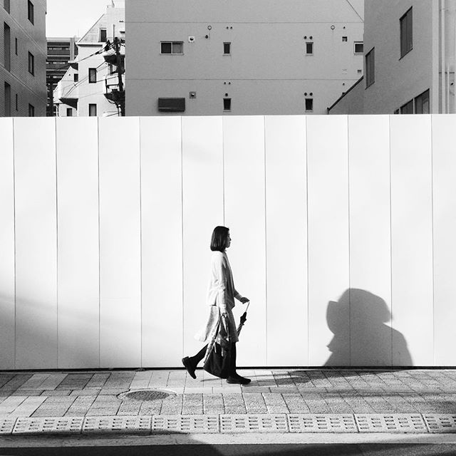 A woman walks along a city street, casting a shadow on a white wall. Black and white.